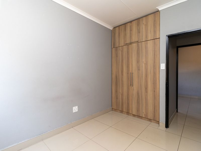 A clean, empty bedroom featuring neutral grey walls, light-toned tiled flooring, and modern wood-finish built-in cupboards.