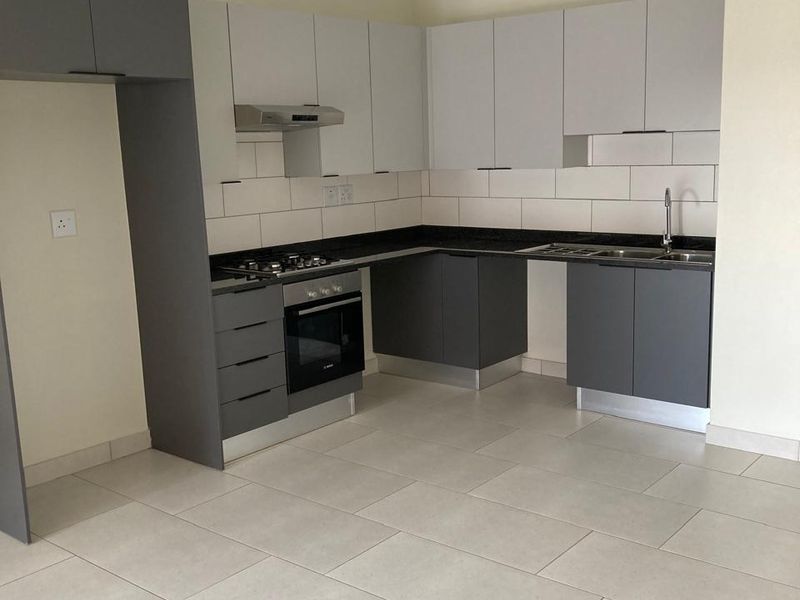 A modern, empty kitchen featuring grey and white cabinetry, stone countertops, built-in oven, gas hob, and neutral tiled flooring.