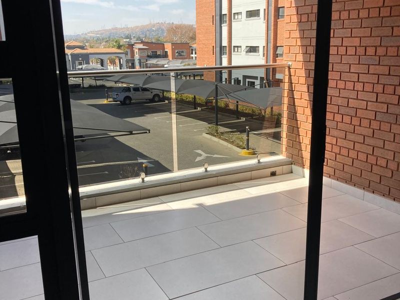 A modern balcony featuring tiled flooring, an exposed brick wall, and a sleek glass balustrade overlooking the complex parking area.
