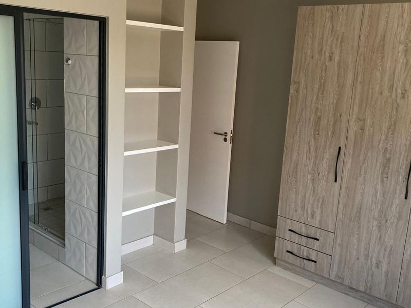 A modern, well-maintained bedroom featuring neutral tiled flooring, built-in wooden wardrobes, integrated shelving, and an en-suite shower with a glass screen.