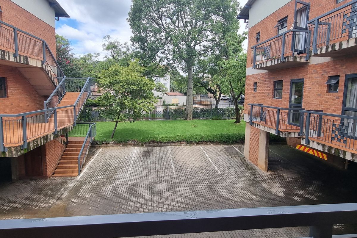 View of the rear exterior of the flat complex, featuring brick facades, metal railings, a paved parking area, and a communal garden space.