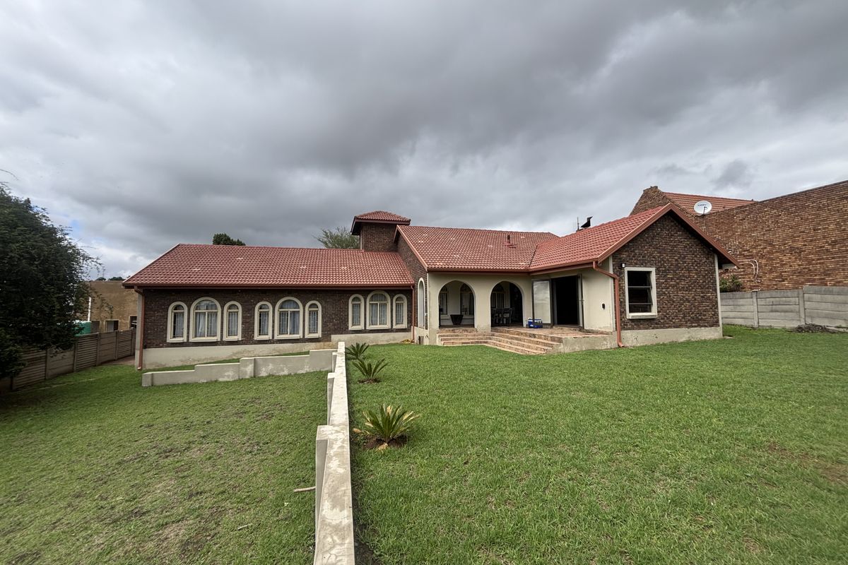 Rear view of a brick house featuring a tiled roof, arched windows, and a covered patio overlooking a spacious lawn with a concrete retaining wall.