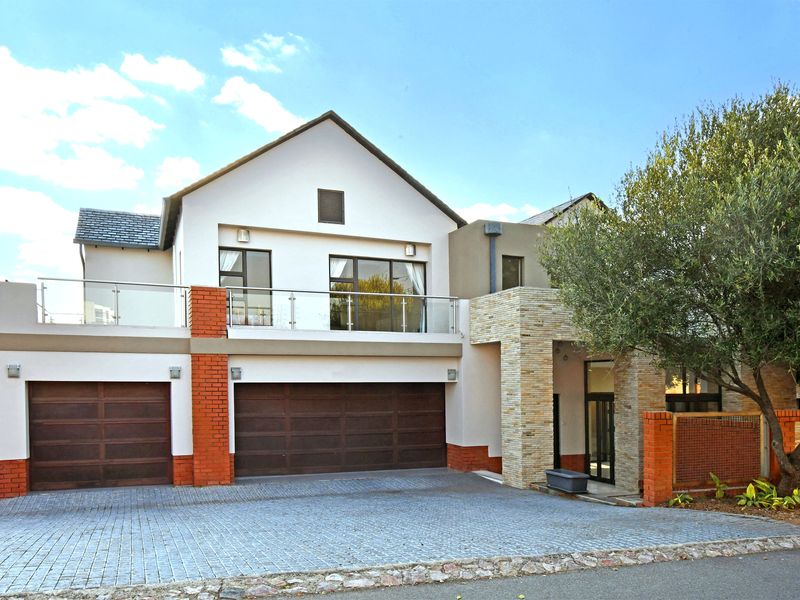 A modern double-story residential facade featuring a double garage, paved driveway, and an upper-level balcony with glass balustrades.