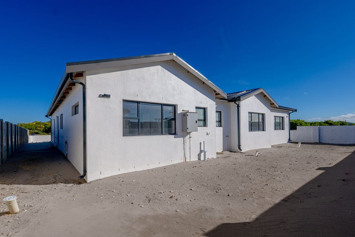 Rear view of the property featuring a white plastered facade, aluminium window frames, and an expansive, undeveloped sandy backyard area.