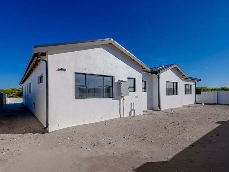 Rear view of the property featuring a white plastered facade, aluminium window frames, and an expansive, undeveloped sandy backyard area.
