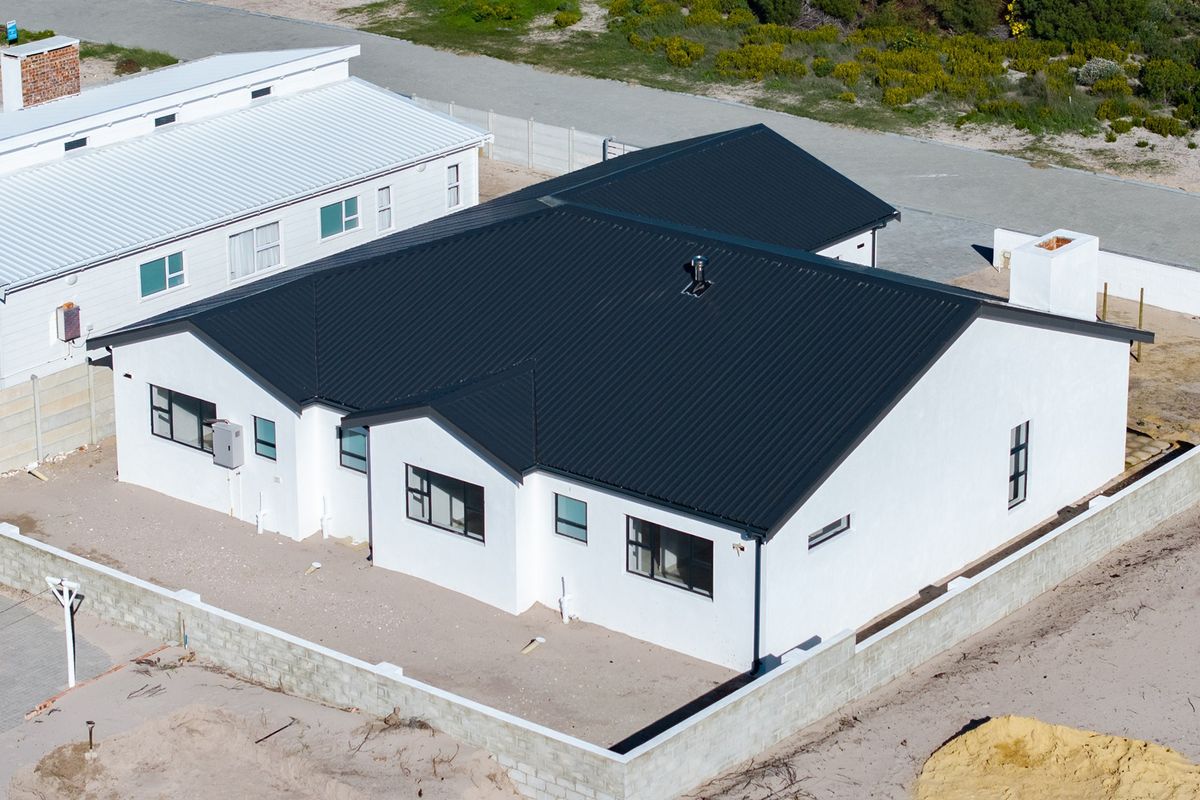 Aerial view of a modern single-story house with a dark corrugated metal roof, white exterior walls, and an unfinished perimeter boundary wall.