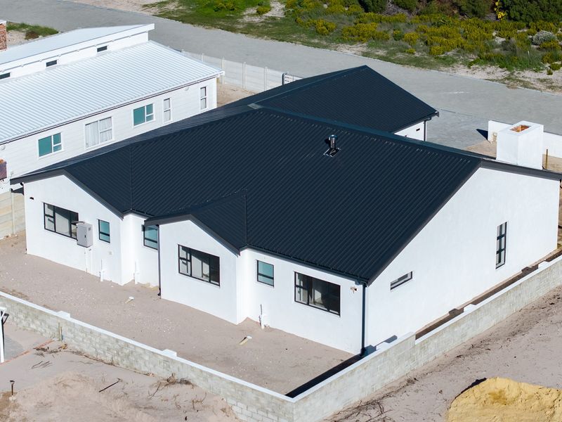 Aerial view of a modern single-story house with a dark corrugated metal roof, white exterior walls, and an unfinished perimeter boundary wall.