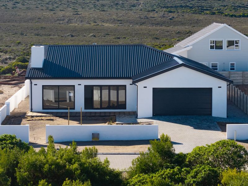 Aerial view of a modern single-story house featuring a dark metal roof, double garage, paved driveway, and white exterior walls in a coastal setting.
