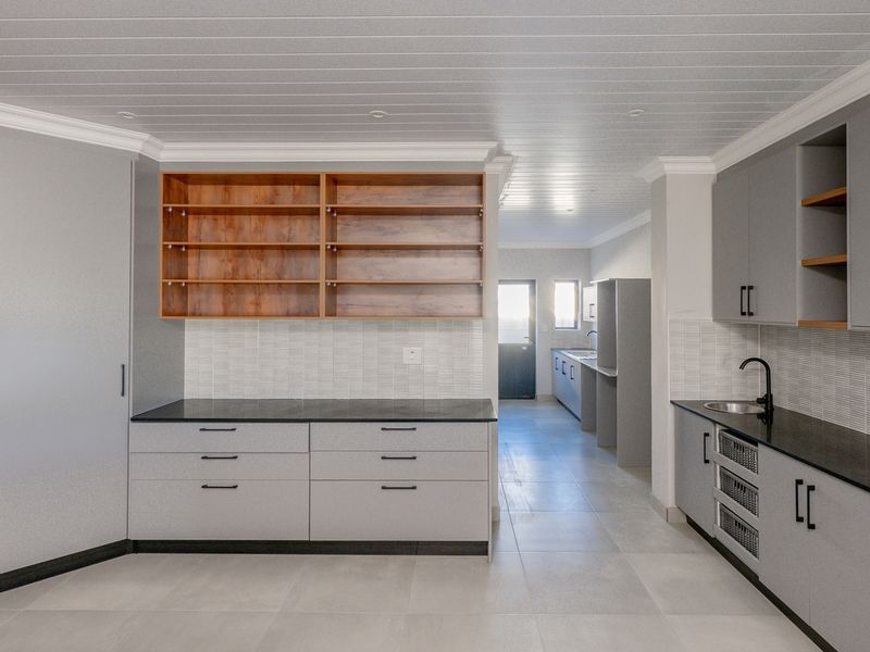 Modern kitchen featuring grey cabinetry, wood-accented open shelving, stone countertops, and a tiled backsplash, with a connected scullery area visible in the background.