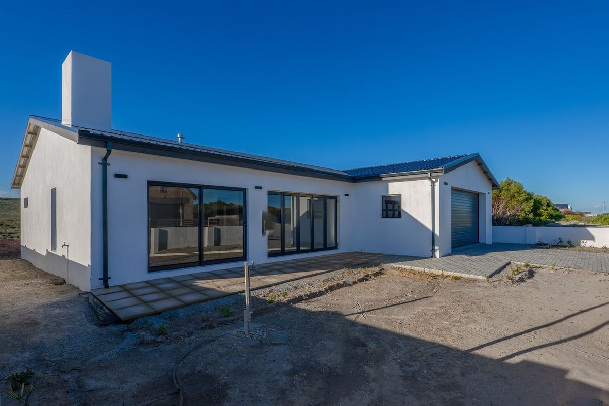 Rear view of a modern white house featuring large sliding glass doors, a paved patio area, and an attached garage with unfinished landscaping.