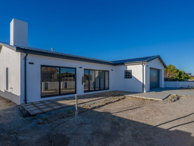 Rear view of a modern white house featuring large sliding glass doors, a paved patio area, and an attached garage with unfinished landscaping.