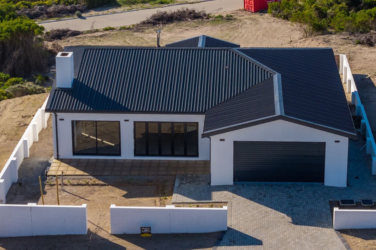 Aerial view of a modern single-story house featuring a dark corrugated metal roof, paved driveway, and a secure boundary wall.