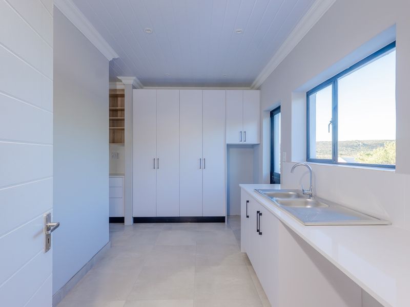 A clean, modern scullery featuring white built-in cabinetry, stone countertops, a stainless steel double sink, and neutral tiled flooring.