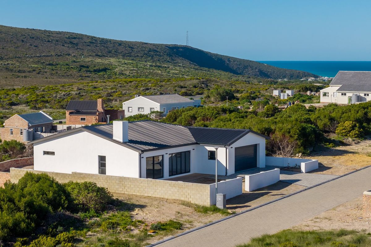 Aerial view of a modern single-story house with a corrugated metal roof, integrated garage, and surrounding brick boundary walls in a coastal landscape.