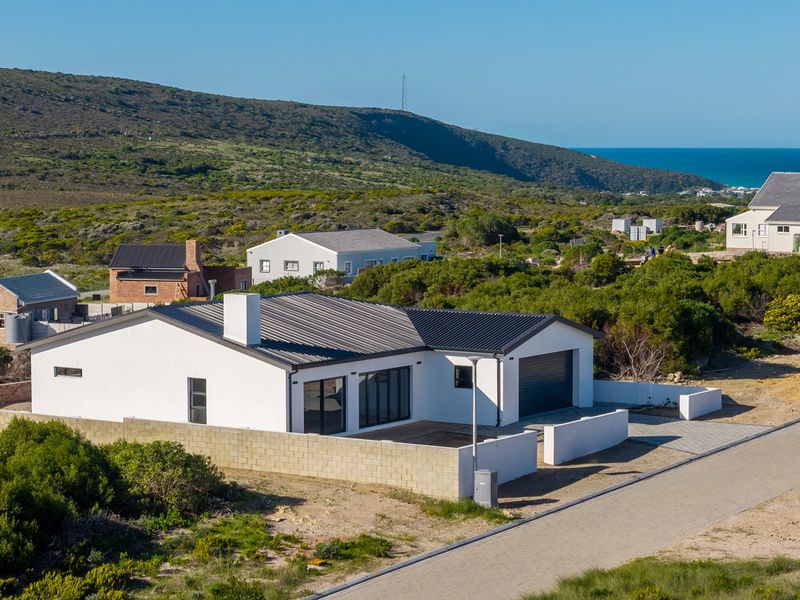 Aerial view of a modern single-story house with a corrugated metal roof, integrated garage, and surrounding brick boundary walls in a coastal landscape.