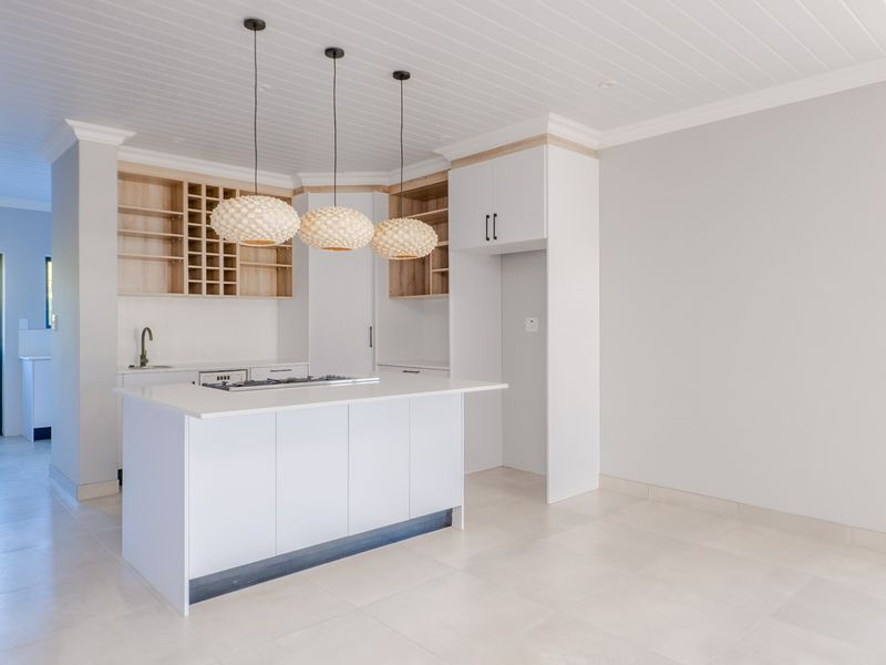 Modern, bright kitchen featuring a central island, clean white cabinetry, light wood shelving, and neutral tiled flooring in an open-plan layout.