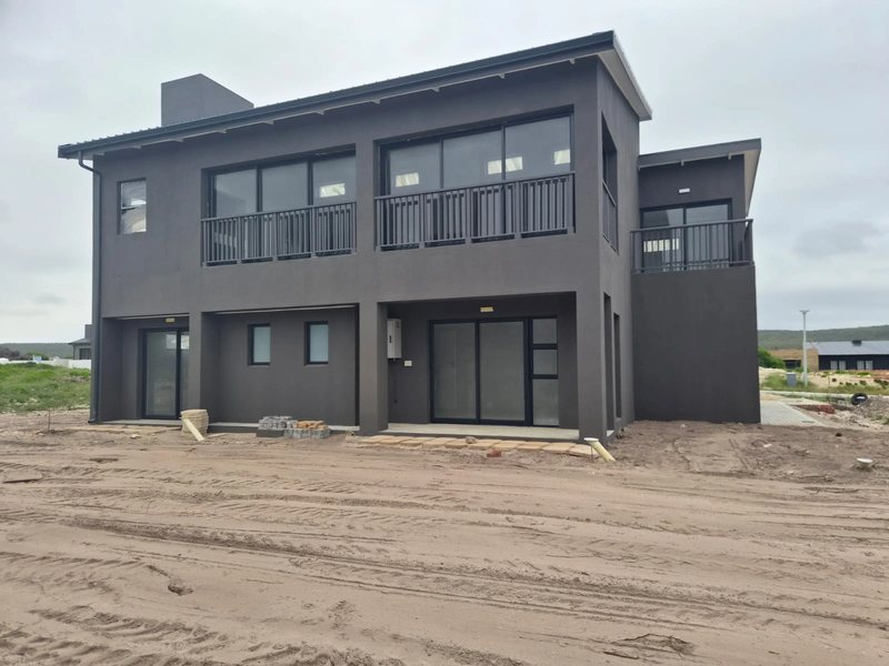 Rear view of a modern two-story house featuring dark exterior finishes, sliding glass doors, and upper-level balconies, currently surrounded by unfinished landscaping.