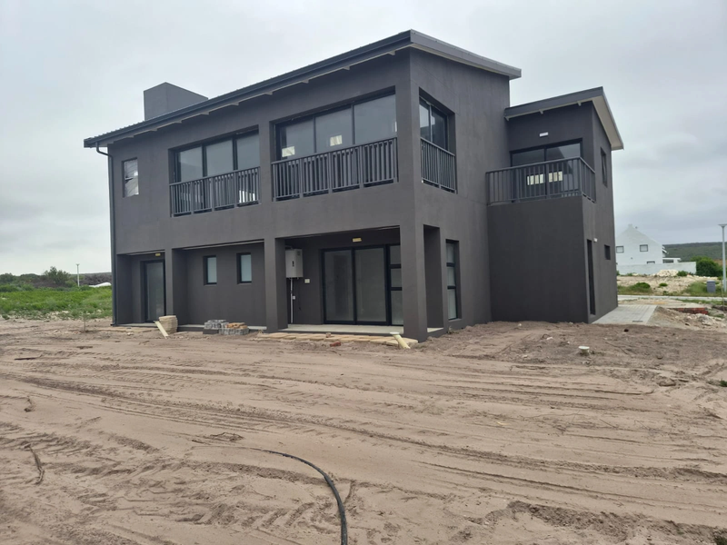Modern double-story house exterior featuring dark grey facade, upper-level balconies, and ground-floor patio area with unfinished surrounding landscaping.