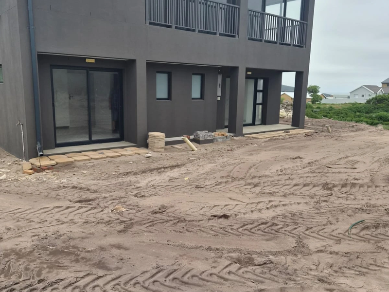 Rear view of a modern two-story house featuring dark exterior cladding, sliding glass doors, and an upper-level balcony, currently undergoing final landscaping.