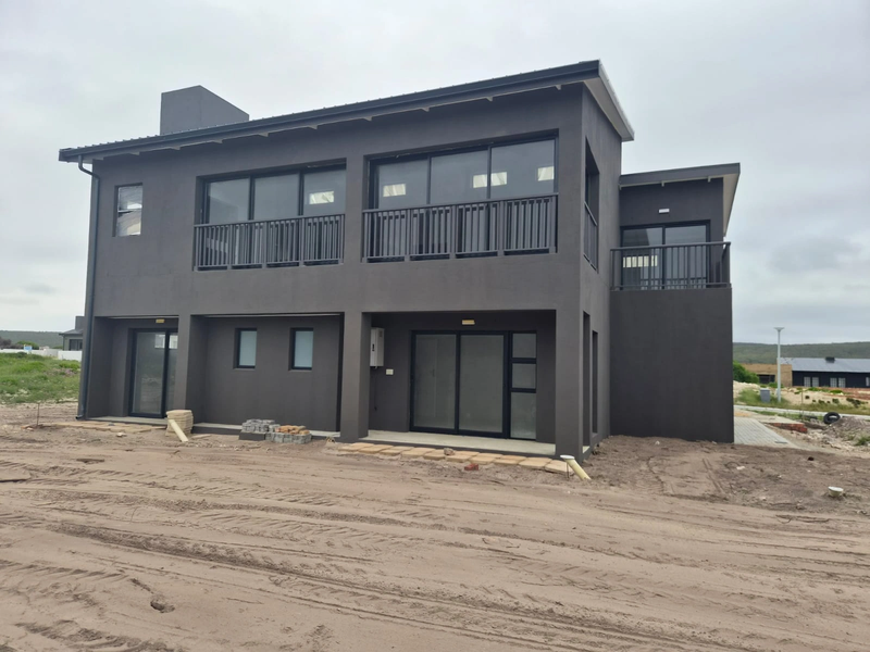 Rear view of a modern two-story house featuring dark exterior finishes, multiple sliding glass doors, and upper-level balconies, currently undergoing final site landscaping.