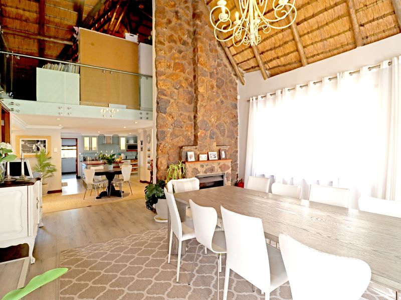 Spacious open-plan living and dining area featuring a prominent stone fireplace, high thatched ceilings with exposed beams, and a mezzanine level with glass balustrades.