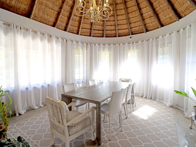 A bright, circular dining room featuring a traditional thatched roof with exposed timber rafters and a central chandelier, surrounded by large windows.