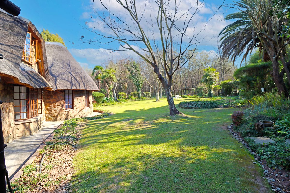 A view of the rear garden featuring a stone-clad house with a traditional thatched roof, surrounded by mature trees and a well-maintained lawn.