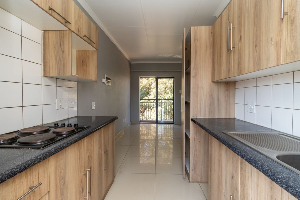 Modern galley-style kitchen featuring light wood-grain cabinetry, dark countertops, electric hob, and tiled flooring, leading to a bright balcony area.