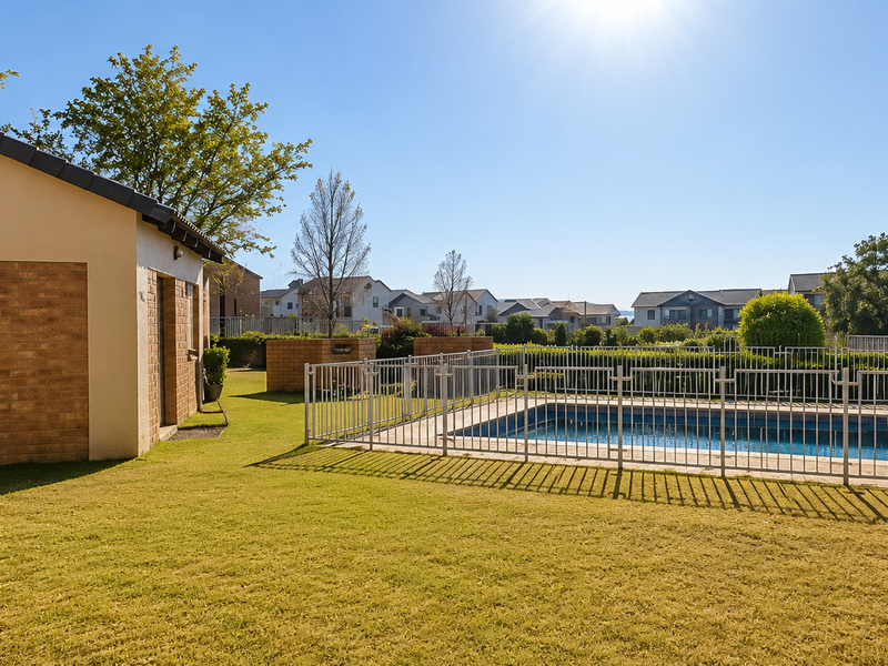 A well-maintained outdoor pool area featuring a fenced swimming pool, manicured lawn, and a brick utility structure within a residential complex.