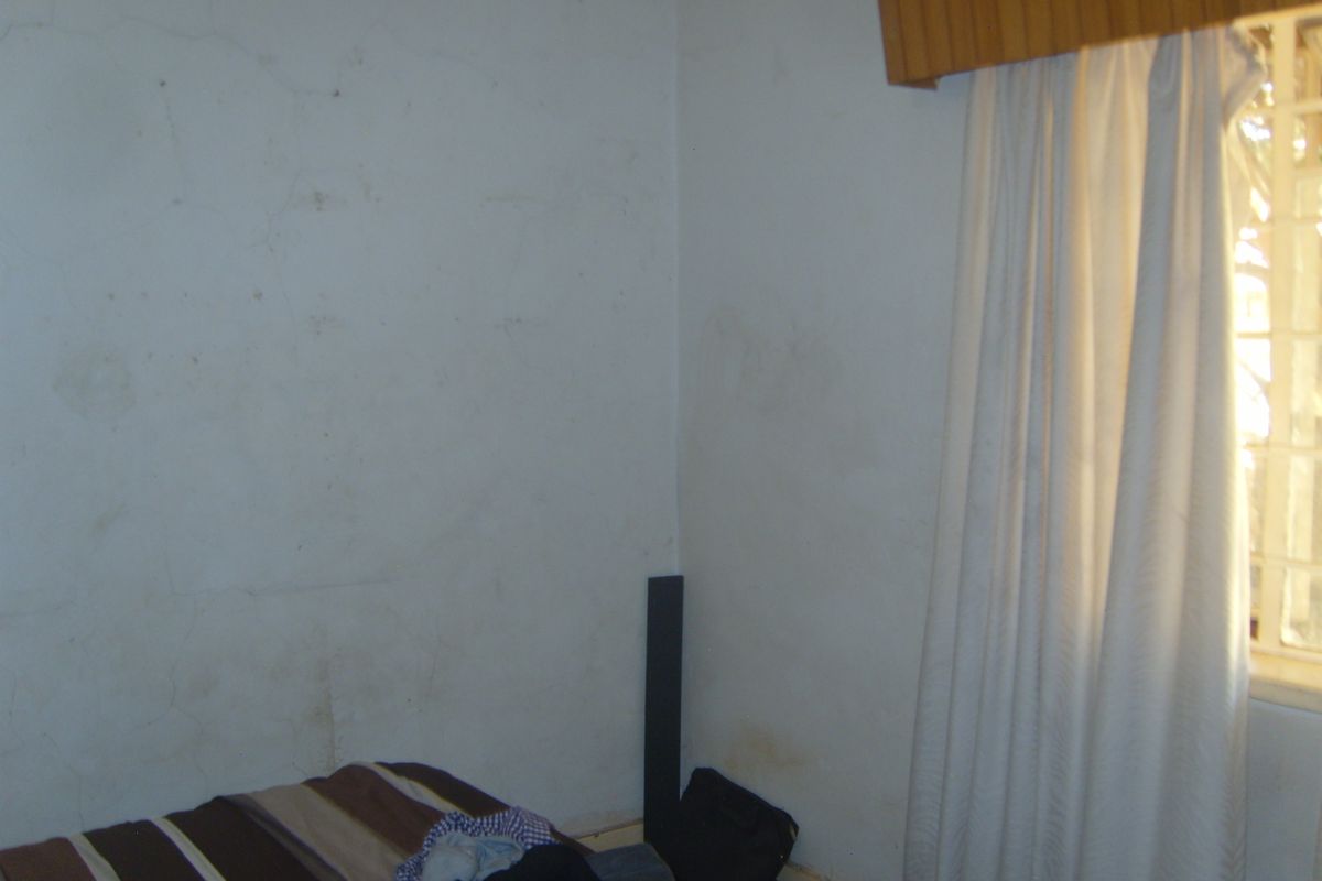 A bedroom featuring a window with security bars and a wooden pelmet, showing significant signs of wear on the wall surfaces.