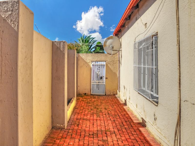 A narrow rear exterior walkway featuring red brick paving, a security gate, and protective window bars along the side of the house.