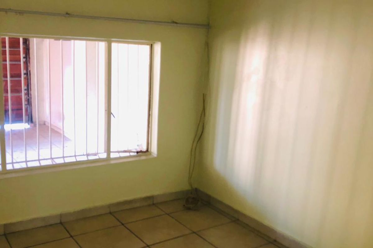 An empty bedroom featuring ceramic floor tiles, security bars on the window, and visible wall imperfections requiring maintenance.