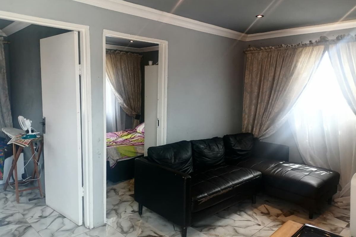 A living room area featuring tiled flooring, recessed downlighting, and crown molding, with an adjacent bedroom visible through the doorway.