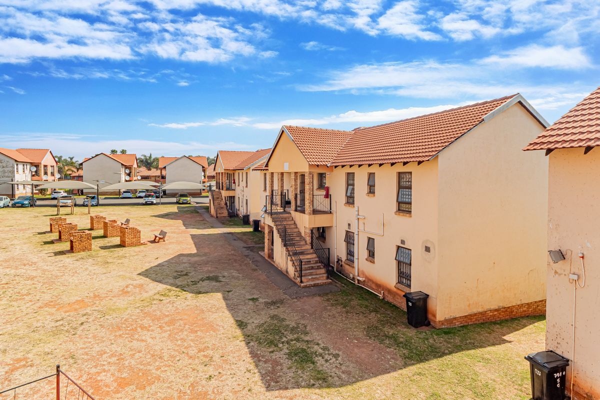 Exterior view of a multi-story apartment complex featuring terracotta tiled roofs, external staircases, and communal brick braai areas in the open yard.