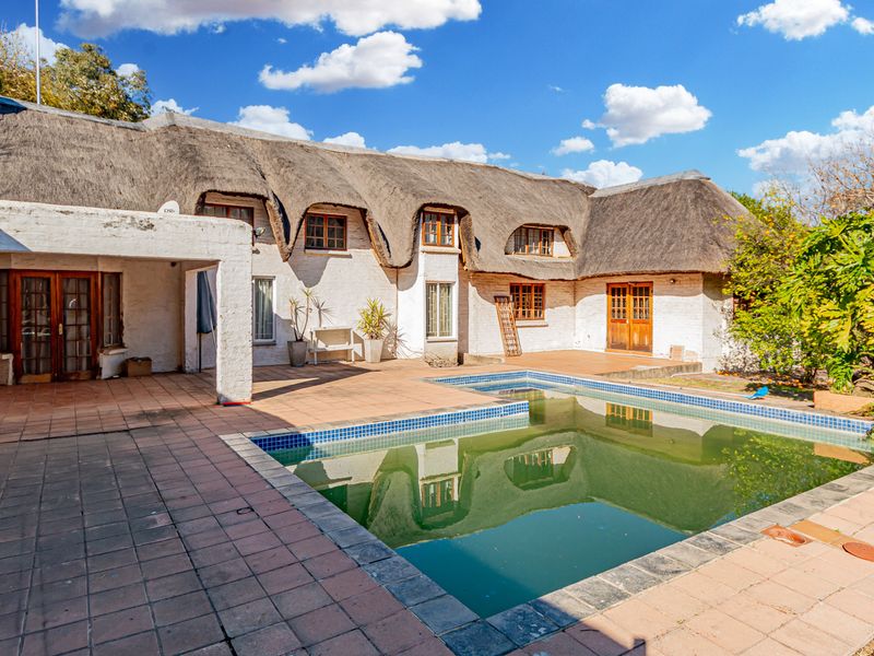 Rear exterior view of a cottage featuring a thatched roof, a tiled patio area, and a swimming pool requiring maintenance.