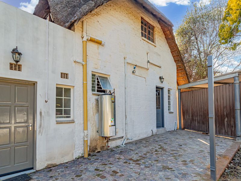 Rear exterior view of a thatched cottage featuring cobblestone paving, an attached carport, and an external electric geyser unit.