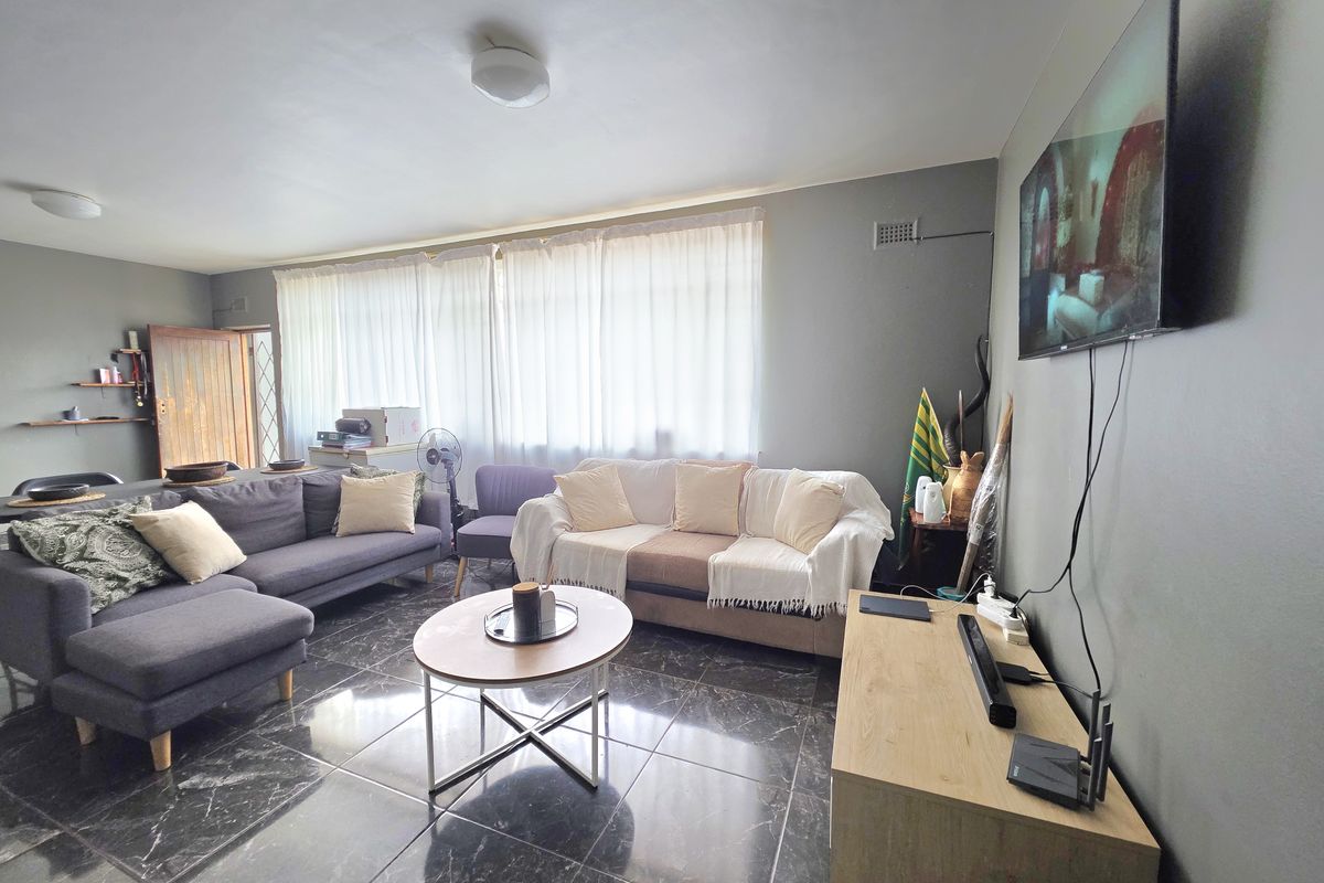 A spacious living room featuring dark marble-look tiled flooring, neutral grey walls, and large windows with sheer curtains, providing a bright and functional living space.