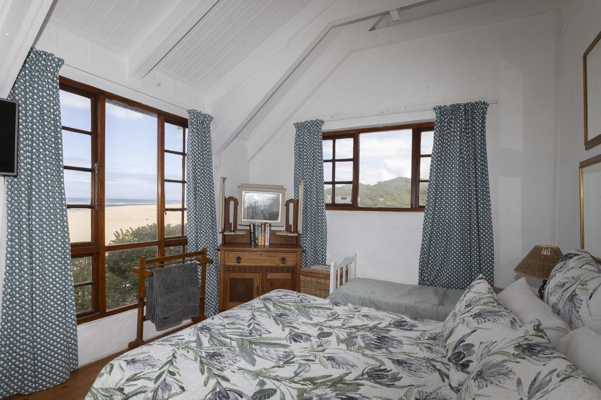 A charming bedroom featuring a vaulted ceiling with exposed beams, timber flooring, and large windows offering scenic coastal and mountain views.