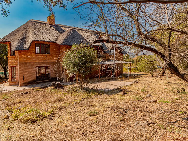 Rear view of a double-story face brick residence featuring a traditional thatched roof, timber windows, and an adjoining paved patio area set in a natural landscape.