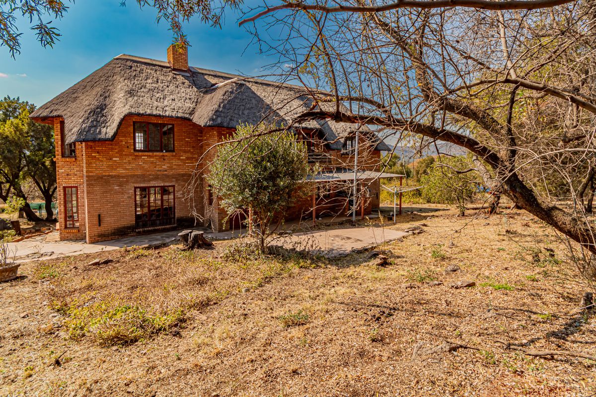 Rear view of a double-story face brick residence featuring a traditional thatched roof, timber windows, and an adjoining paved patio area set in a natural landscape.
