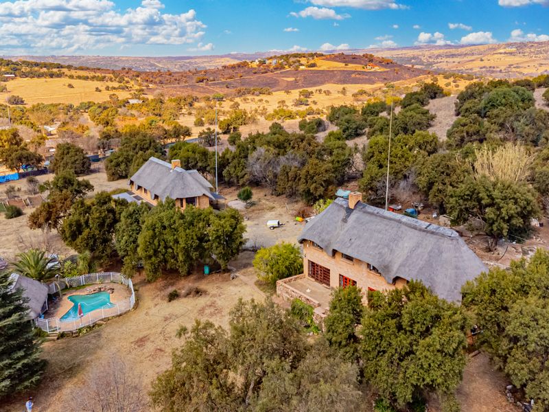 Aerial view of a small holding property featuring two thatched-roof dwellings, a swimming pool, and expansive natural landscape.