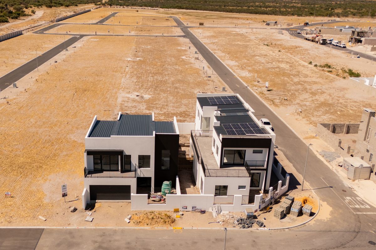 Aerial view of two modern double-story residential properties under construction, featuring solar panels, metal roofing, and surrounding development infrastructure.