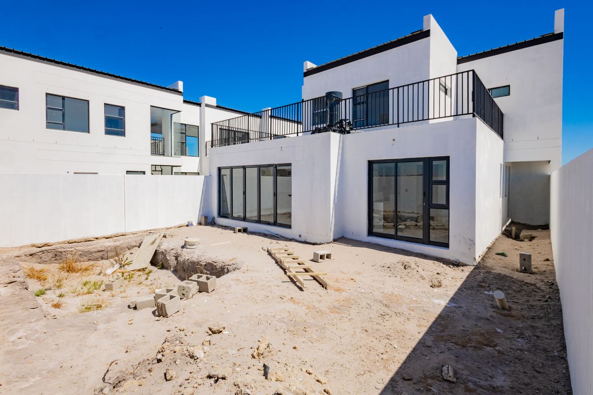 Rear view of a modern two-story house featuring a balcony with metal railings, large sliding glass doors, and an unfinished backyard area.