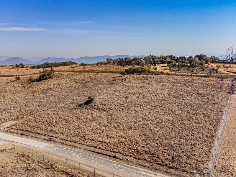 An aerial view of a large, vacant residential plot featuring natural grassland, perimeter fencing, and direct access to a paved road.