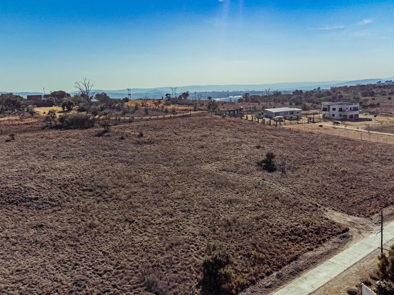 An aerial view of a large, undeveloped residential plot featuring dry vegetation, perimeter fencing, and adjacent paved road access.