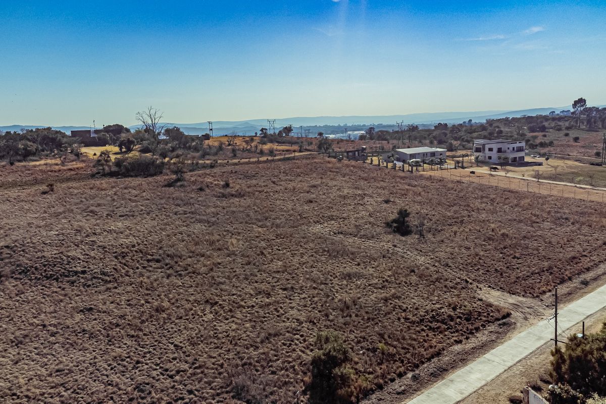An aerial view of a large, undeveloped residential plot featuring dry vegetation, perimeter fencing, and adjacent paved road access.