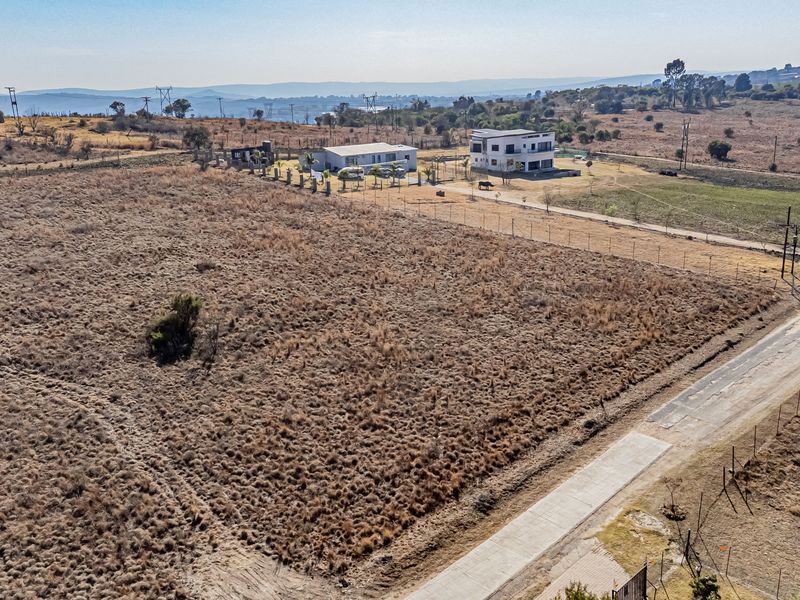 Aerial view of a vacant residential plot featuring perimeter fencing and adjacent paved road access in a developing rural area.