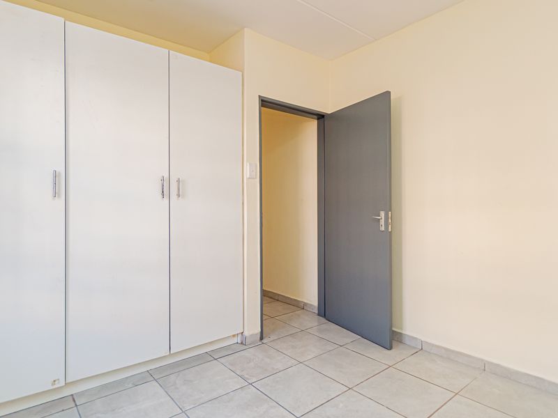 A clean, empty bedroom featuring built-in white cupboards, neutral wall finishes, and tiled flooring with access to an adjacent room.