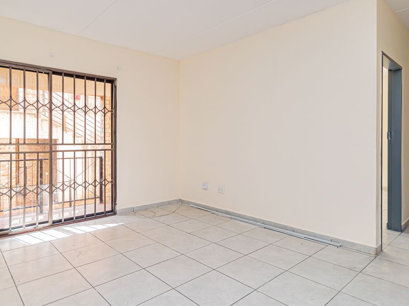 A bright, empty living room featuring neutral tiled flooring, a secure sliding door with metal security gate, and access to an adjacent hallway.