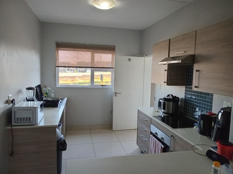A functional kitchen featuring modern wood-finish cabinetry, integrated appliances including an oven and hob, tiled flooring, and a window with venetian blinds.